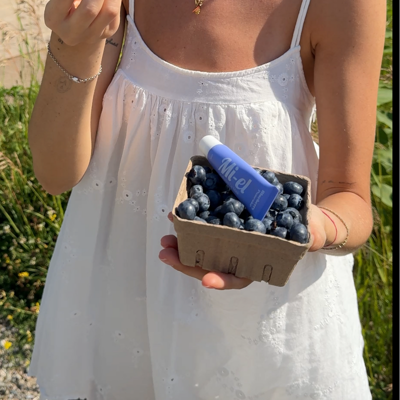 A woman outdoors wearing a white spaghetti-strap dress and holding a small container of blueberries with a blue tube, standing in a grassy field. Blueberry pancake lip mask